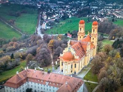 This aerial, slightly blurry image depicts an expansive, historic complex of buildings nestled within a landscape of rolling hills, green fields, and autumnal trees. The central focus is a large, ornate, yellow church with multiple red-domed towers and a prominent central dome. Adjacent to the church is a long, multi-story building with a red-tiled roof and numerous windows. In the distance, a town with houses and a church spire is visible, set against a backdrop of darker, wooded hills. The overall scene suggests a rural or semi-rural European setting, possibly during late autumn or early spring, given the bare trees and muted colors of the foliage. The lighting indicates it is daytime, though the overcast sky contributes to the somber atmosphere. There is no discernible activity or people in the image, focusing solely on the architectural and natural landscape.
