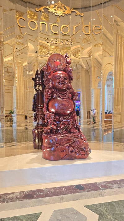 In the lobby of Concorde Casinos in Agios Epiktitos, Northern Cyprus, a large, intricately carved red wooden statue of a laughing Buddha stands prominently on a raised marble platform. The statue is adorned with a beaded necklace and a circular emblem with a red inset on its head. Behind the statue, mirrored walls reflect the opulent interior, revealing ornate golden architectural details, arches, and columns. In the background, a few individuals can be seen, some walking, and others possibly seated, blurred by the reflections and distance. The floor is a polished marble with geometric patterns. The scene suggests a luxurious and grand establishment.
