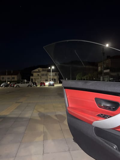 The image captures a nighttime scene from inside a car parked in Zverino, Bulgaria. The open car door reveals a black and red interior, with the red being the leather upholstery of the door panel. The car is parked on a paved square, illuminated by a streetlamp visible in the background. Several cars are parked in a row across the square, with buildings lining the street behind them. The sky is dark and clear, with a small light visible in the upper left corner, possibly a distant star or airplane. The overall impression is one of a quiet, possibly residential, area at night.