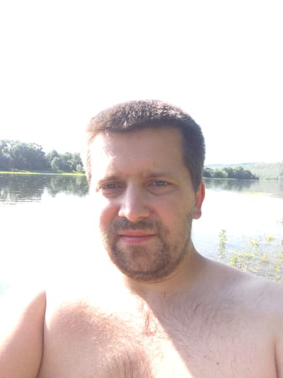 A man is taking a selfie while in the water, possibly swimming or wading. The image is taken from a low angle, with the man's chest, shoulder, and part of his face visible. He has fair skin, light brown hair, and a beard. His eyes are open and looking towards the camera. The background shows a body of water with reflections of trees and the sky, suggesting a sunny day. The man's expression appears neutral or relaxed. The scene is outdoors, with vegetation visible along the water's edge. The photograph appears to be taken during daylight hours, likely in the late morning or afternoon due to the brightness of the sky and the shadows cast. The provided location context of Zalishchyky, Ukraine, suggests a rural or natural setting. There is no visible text in the image.