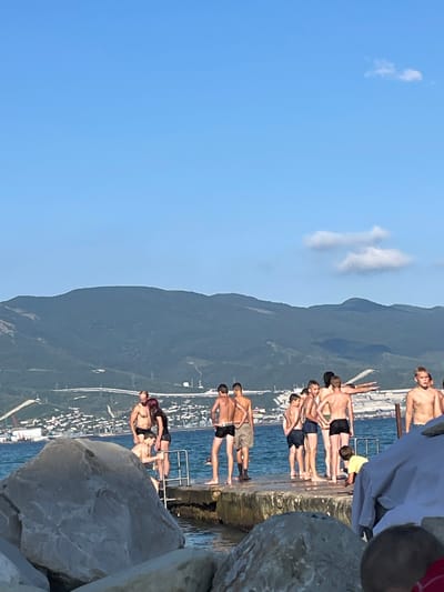 A group of individuals, mostly young males, are gathered on a concrete pier by the sea. They are dressed in swimwear. Some are standing, looking out towards the water or interacting with each other, while others appear to be preparing to enter the water. In the background, a mountainous landscape with a coastal city settlement is visible. The sky is clear blue with a few scattered clouds. The scene appears to be set in the city of Ol'ginskii, Russia, likely during a warm weather season.