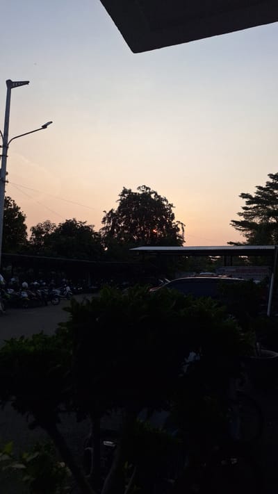 The image depicts an outdoor scene at dusk in Karawang, Indonesia. The sky is a gradient of light blue to orange near the horizon, where the sun is partially obscured by a tree. Silhouetted against the sky are trees, utility poles, and the corner of a building. Below the tree line, a parking area is visible with several motorcycles parked closely together. Vegetation borders the parking area on the right.
