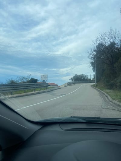 A road ascends slightly, marked with a central white line. Guardrails flank the roadside, and a sign indicating "Oliva Villages" is visible. The sky is blue, adorned with streaky clouds. Trees border the opposite side of the road. The photograph appears to be taken from within a car. The vehicle's dashboard and interior are partially visible in the foreground.