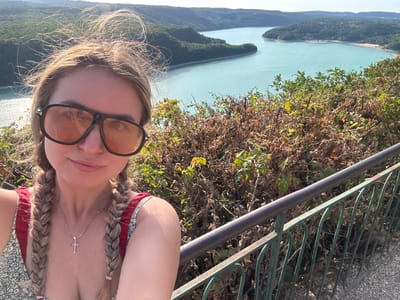 A young woman with blonde hair styled in two braids is taking a selfie outdoors. She is wearing large, dark-rimmed sunglasses with orange lenses, a light-colored dress with a floral pattern, and a red top with thin straps. A silver cross necklace hangs around her neck. The background features a body of water with green, tree-covered hills surrounding it, a scenic landscape that is likely located near Moirans-en-Montagne, France. A dark metal railing is visible in the foreground to the right of the woman. The lighting suggests it is a sunny day.