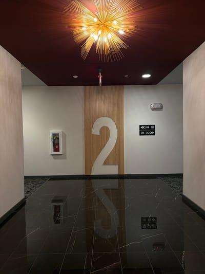 The image shows a hallway with a large number "2" on the wall, made of wooden slats. A fire extinguisher is mounted on the left wall. A directional sign with room numbers is on the right wall. The floor is highly polished black marble, reflecting the ceiling light fixture, which is a starburst design with multiple bulbs. The ceiling is a dark red color. The location is Maryland Heights, United States of America, likely in a hotel or apartment building. There are no people visible in the image.