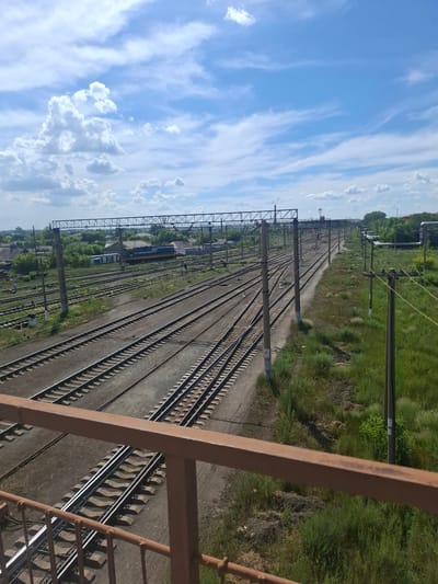 The image depicts a railway yard with multiple tracks stretching into the distance. The perspective is from an elevated vantage point, likely a bridge or overpass, as a rusty brown railing is visible in the foreground. The sky is a vibrant blue with scattered white clouds, indicating a clear and sunny day. Green grass and trees line the sides of the tracks, suggesting a rural or semi-urban setting. Several utility poles with overhead wires are also present, indicating the presence of an active railway line. In the mid-ground, a train with a dark blue and yellow body is partially visible on one of the tracks. The overall scene conveys a sense of industrial activity and travel. The time of day appears to be daytime, given the bright sunlight and clear sky. No text is visible in the image. The location context is Atbasar, Kazakhstan.