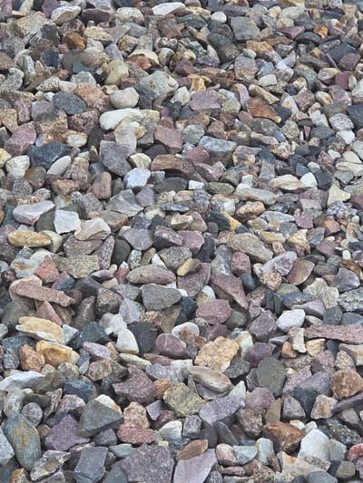 The image displays a close-up, overhead view of a pile of gravel. The gravel consists of various rock fragments, ranging in size from small pebbles to larger chunks. The rocks exhibit a diverse color palette, including shades of grey, brown, tan, white, and hints of purple and pink. Many of the stones are angular and irregularly shaped, suggesting they have been crushed or broken. The texture appears rough and stony. The lighting is consistent and even, indicating it is likely daytime with diffused light, possibly overcast. There are no people or specific objects other than the gravel itself, and no discernible text is present in the image. The scene is a static arrangement of stones, lacking any action or event. Without further context, the specific location cue of Frolovo, Russia, cannot be visually confirmed from the image content alone.