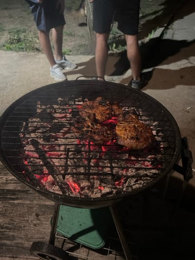 An outdoor, dimly lit scene in Stolnik, Bulgaria, captures a circular charcoal grill with three pieces of browned meat cooking on its grate. Glowing red embers and grey ash are visible beneath the meat. The grill's support structure, including a wheel and a green plastic tray, is partially visible in the lower-left foreground. In the background, the lower legs of two individuals are visible on a concrete surface: one person wears blue shorts and white sneakers, the other wears dark shorts and dark shoes. Distinct shadows are cast from their figures onto the concrete. Sparse vegetation is present in the far background, and a cigarette butt lies on the concrete surface near the right individual's foot.