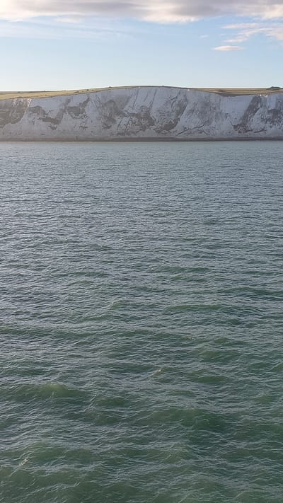 The image shows a panoramic view of the White Cliffs of Dover in the United Kingdom. The cliffs are a prominent natural landmark on the English coastline. The image captures the stark white chalk cliffs rising from the sea. The sea itself is a deep blue-green, with gentle waves creating a textured surface. Above the cliffs, a strip of green grass runs along the clifftop. The sky above is a pale blue, with a few wispy clouds. The lighting suggests it is either early morning or late afternoon, with soft sunlight illuminating the scene. There are no people or boats visible in the image.