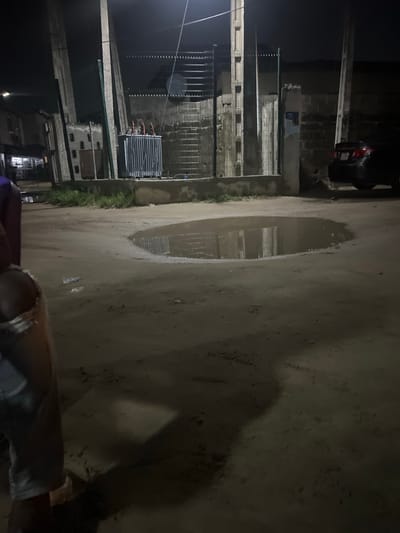 The image captures a dark, unpaved street or path in Isunba, Nigeria, at night. A large, prominent puddle of water occupies the center of the ground, reflecting the structures lining the path and creating an impression of recent rain.

On the left side of the path, a series of unpainted or weathered buildings with block walls and visible concrete pillars stand. Metal fencing and vertical bars are integrated into the facade of these structures. A piece of industrial equipment, possibly a transformer or air conditioning unit with visible piping, is mounted on one of the buildings. Towards the upper part of the image, the rear end of a dark-colored parked car is partially visible.

The ground is rough, suggesting it is unpaved, and appears damp around the puddle. Scattered debris or litter is faintly visible on the ground. Along the edge of the path near the buildings, some green grass or vegetation can be seen.

The scene is dimly lit, indicating sparse artificial light sources. The perspective seems to be from a person on a motorcycle or scooter, as part of a leg and clothing are visible in the bottom right corner of the frame. There is no discernible text visible within the image.