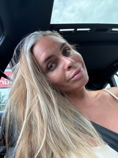 A woman with blonde hair and blue eyes is in the car. The car appears to be parked or stopped in Tha Chana, Thailand, indicated by the Thai signage in the background. The woman is looking at the camera and smiling. She is wearing a white tank top. The sunroof of the car is open, and daylight is visible.