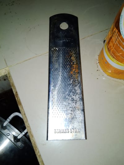 The image shows a rusted stainless steel grater lying on a tiled floor. To the right of the grater is a yellow plastic container, possibly for food or spices. A portion of a metal pot with a clamp is visible in the bottom left corner. The floor appears to be dirty with some debris scattered around. The scene is indoors, likely in a kitchen or storage area. There are no people visible in the image. The location is identified as Sendangrejo, Indonesia.