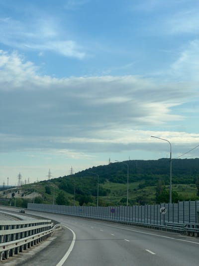 The image depicts a highway curving alongside a hillside with a mix of trees and grassy areas. The highway has a gray noise barrier along one side and a white line marking the edge of the lane. Streetlights line the road. In the background, high-voltage power lines are visible. A speed limit sign of 60 is posted. The sky is cloudy with patches of blue. The location is likely Inkerman, Ukraine.