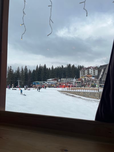 The image captures an active winter ski resort scene in Stoikite, Bulgaria, viewed from an interior window framed by a wooden sill and adorned with suspended, illuminated string lights. The sky is heavily overcast with grey clouds, indicating diffuse daylight conditions.

In the foreground and midground, a broad, snow-covered ski slope is populated by approximately 20-30 individuals engaged in recreational activities. People in winter clothing are seen skiing, standing, and walking across the snow. One individual in a blue jacket is kneeling beside a snowboard lying on the snow in the lower-left quadrant. To the left, a ski lift ascends into a dense coniferous forest that forms a natural backdrop across the middle of the scene.

Beyond the forest and at the top of the ski slope, a cluster of resort buildings is visible. This includes a prominent multi-story hotel-style structure with white and brown facades. Adjacent to it are various smaller commercial or leisure buildings, some featuring colorful elements and Cyrillic signage, such as "ОСВ." A retaining wall, possibly concrete or stone, topped with a railing, separates the elevated area with the buildings from the main ski slope. Fencing and other resort infrastructure are also present near the buildings.