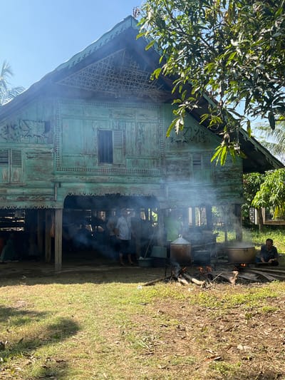 In Pulo, Indonesia, a traditional wooden stilt house with weathered turquoise paint is the central focus of the scene. The house has intricate carvings and patterned latticework on its upper sections and features louvered windows. Smoke billows from an outdoor cooking area in front of the house, where large metal pots are situated over open fires. Several individuals are present; some are gathered under the stilt house, while others are tending to the fires and pots. The ground is covered in grass and dry leaves, and lush green trees are visible in the background, suggesting a rural or semi-rural environment. The sky is clear and blue.