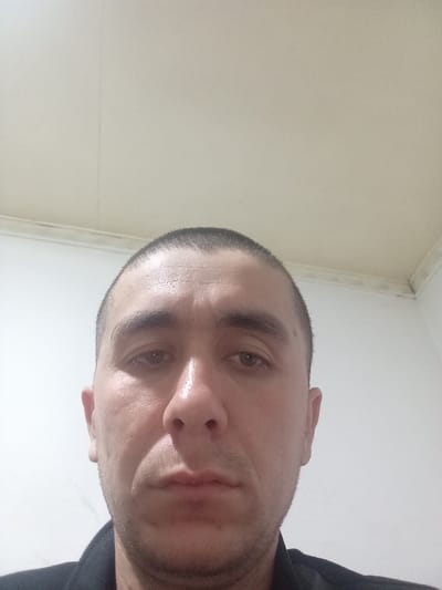 A close-up selfie of a man with a very short, buzzed haircut. He has fair skin, dark eyebrows, and a serious expression. His eyes are open, and his nose is prominent. He's wearing a dark-colored shirt or jacket. The background consists of a white wall and a textured ceiling, with a slightly visible corner in the upper right. The lighting appears to be indoor and somewhat diffused.
