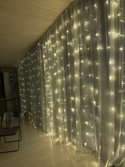 An interior shot shows a room with a wooden ceiling and floor. A long white sheer curtain is draped across the right side of the frame, illuminated by numerous small, warm-toned fairy lights. The lights are arranged in vertical strands, creating a sparkling effect against the fabric. To the left of the curtain, a refrigerator is visible, and a dark metal folding chair is placed in the foreground. A light blue cloth is draped over the chair. The overall impression is one of a cozy, dimly lit space with decorative lighting. The image does not depict any specific landmarks or features that would pinpoint its location in Starye Chernitsy, Russia, nor are there any people or discernible actions taking place.