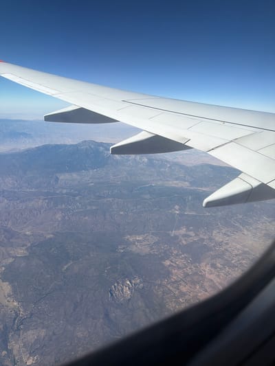 The image, taken from an airplane window, shows a vast, arid landscape with mountains in the distance under a clear blue sky. An airplane wing dominates the upper portion of the frame, casting a shadow over the terrain below. The ground is a patchwork of brown and green, with evidence of human settlement, including scattered buildings and roads, visible in the lower half of the image. The scene suggests a flight over a rural or mountainous region, not within the city of Houston, Texas.