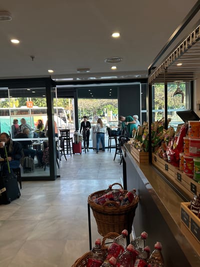 The scene takes place inside a cafe located in Granadilla de Abona, Spain, at coordinates 28.048, -16.578. Several people are seated at tables near a large window that overlooks a street, where a public bus is parked. A group of people are standing near the window, some of them looking outside. The cafe is clean and modern, with a tiled floor and exposed ceiling beams. On a counter in the foreground are multiple baskets filled with bottled beverages, and a rack behind it is stocked with bags of chips. The cafe is well-lit by overhead lights.