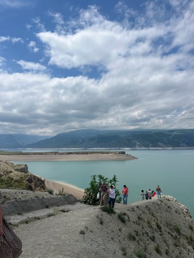 A scenic outdoor shot, rotated counterclockwise, features a cliff edge overlooking a reservoir with several individuals. The sky occupies a significant portion of the left side, displaying a blend of blue and cloud cover.
On the right, a rocky cliff edge serves as a viewpoint, with a group of people standing near the edge, looking out over the landscape. Visible on the cliff, nearest the water's edge, is a tree. Additional individuals are standing further away along the cliff.
The reservoir fills the lower part of the image, exhibiting a light turquoise color, and is flanked by mountain ranges in the distance. A section of the shoreline is visible, appearing as a narrow sandy strip between the water and the mountains.
The scene is likely taken in or around Chirkey, Russia, as indicated by the presence of Chirkey Reservoir.