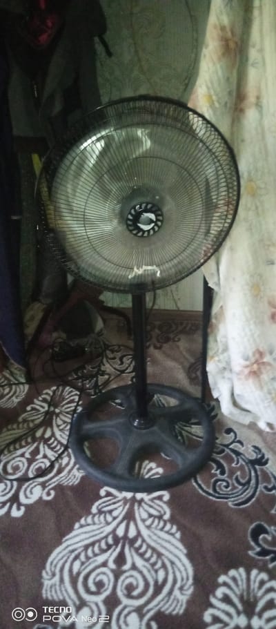 A standing fan with a black base and stand is positioned in the center of the frame, facing slightly to the left. Its circular grill is made of thin metal wires, and the blades within are visible, suggesting it may be in operation. The fan rests on a patterned rug with a brown background and intricate white and black designs. Behind the fan, a light-colored curtain with a floral pattern is visible to the right, and to the left, darker fabric, possibly clothing or a drape, hangs down. The scene appears to be indoors, in what could be a domestic setting in the city of Tahmach, Uzbekistan.