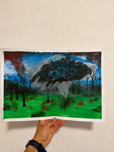 The image is a photograph of a painting held by a hand against a textured white wall. The painting depicts a landscape with a dark, abstract shape dominating the center, surrounded by bare trees and a green, grassy ground. The sky is a mix of blue and gray with streaks of red. The overall impression is one of desolation or a chaotic event, possibly a storm or a fire. The hand holding the painting has a dark watch on its wrist. The location in Nienburg/Weser, Germany is not discernible from the image itself.