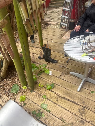 This image depicts an outdoor setting in Romainville, France, characterized by a weathered wooden deck. On the left, several tall, green wooden or bamboo poles rise from a bed of dried leaves and support hanging wind chime components, including hollow bamboo sections and dried leaves on strings. A pair of black cowboy boots, featuring intricate stitching and textured material, stands upright on the wooden deck. Green foliage, resembling ivy, grows on and between the deck boards.

To the right, a round, silver-colored metal table with a striped surface and a central pedestal base is present. Arranged on the table are a white power adapter with attached cables, a small black electronic device, a white rectangular container with blue markings, a rusted multi-pronged metal gardening tool, a yellow-green clothes peg, and a pair of orange-handled pruning shears. A small black cylindrical object lies on the deck near the table's base.

A person is partially visible on the right, seated at the table, wearing a dark jacket and dark pants, with hands resting on their lap. In the background, behind the poles on the left, the lower legs and dark rain boots of a second person are visible. Beyond them, a stone pathway with steps ascends toward a red-painted structure, against which a metal step ladder is propped.