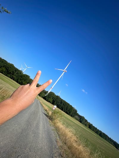 A first-person perspective shot shows a hand with two fingers extended in a peace sign, as if it's about to be photographed while walking on a gravel path in Berfa, Germany. A vast, clear blue sky dominates the upper portion of the frame. In the middle ground, a field of dry grass and a green meadow stretch towards a line of dense trees bordering the landscape. Several wind turbines are visible in the distance, with white blades and red tips, dotted across the horizon. One prominent turbine is positioned closer to the right side of the image, its blade angled upwards. A road sign, possibly indicating a bike path, is situated on the right side of the gravel path, near the edge of the meadow. The scene is illuminated by bright sunlight, casting shadows and highlighting the textures of the gravel path and dry grass.