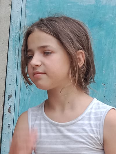 This is a close-up, medium shot of a young girl with shoulder-length brown hair, facing left with her head slightly turned. She has light skin and her expression appears neutral or slightly contemplative. She is wearing a light gray sleeveless shirt with thin white stripes.
The background consists of a distressed, weathered blue wooden surface, possibly a door or wall, with patches of peeling paint. To the left of the blue surface, there is a section of a rough, light-colored wall, likely concrete or stucco, also showing signs of wear.
The lighting suggests it is daytime, likely outdoors. The overall scene is casual and appears to be in a somewhat rural or older urban setting. There is no visible text in the image. The girl's hair is slightly windswept, suggesting a gentle breeze.