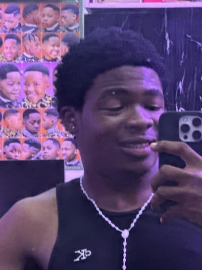 A person with dark skin and short, curly black hair is taking a selfie. They are wearing a black shirt with a white logo on the upper right. A white beaded lanyard hangs around their neck. They are holding a black smartphone with multiple camera lenses in their hand. Behind the person is a wall covered with a repeating pattern of images of a man's face. A marble-like surface is visible in the upper portion of the frame. There is no information available in the image that identifies the specific location as Ogwashi-Uku, Nigeria.