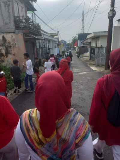 The image displays an outdoor scene on a paved asphalt street in Bojongherang, Indonesia, under an overcast sky. A procession of approximately 15-20 individuals is moving away from the camera, along the street.
In the immediate foreground, three individuals are partially visible from behind. The leftmost figure wears a red top and white lower garment. The central figure, closest to the camera, wears a white top with a multi-colored patterned hijab/scarf overlayed by a red hijab. The rightmost figure wears a red top, red hijab, and a black backpack.
Ahead of these foreground individuals, a line of approximately 10-15 people, predominantly younger males, walks in the same direction. One youth wears a white t-shirt with the number "90" on the back. Another youth on the left side of the line is seen holding a white bottle or object.
The street is flanked by buildings. On the left, an older, two-story building with a weathered, light-colored facade and a small metal-railed balcony on the second floor is visible, with green vegetation at its base. On the right, newer-looking, light-colored residential buildings with fences and gates line the street.
Numerous overhead utility wires are visible against the sky. Multiple utility poles are present, and a tall cellular communication tower is prominent in the distant background. A red and white Indonesian flag is displayed on a pole further down the street on the right. In the distance, a person on a scooter and a parked white car are visible.