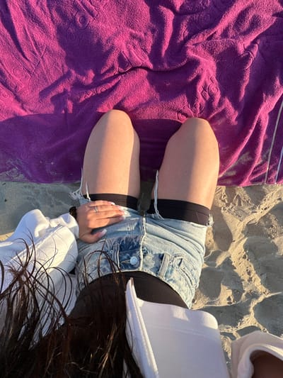 An overhead, first-person view captures an individual reclining on a sandy beach in Manhattan Beach, United States. The person is dressed in light blue distressed denim shorts worn over black undergarments, with a white long-sleeved top partially visible. Their left hand, adorned with a silver ring on the ring finger and manicured nails with white tips, rests on the right thigh. Dark brown hair extends into the top left of the frame. A crumpled magenta beach towel with an embossed pattern lies on the sand above the individual's legs. The scene is illuminated by direct sunlight, casting clear shadows on the sand.