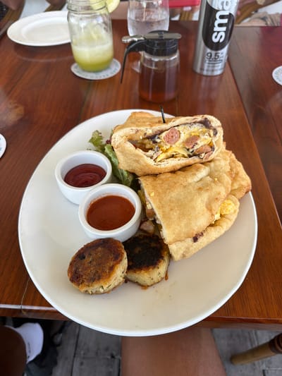 A meal is presented on a wooden table in an outdoor dining environment within San Pedro Town, Belize. A white oval plate holds a large, deep-fried bread pocket, sliced open to reveal scrambled eggs, sliced hot dogs, and black beans. Two small white ramekins contain red ketchup and a darker red sauce. A garnish of green lettuce is present. Three pan-fried round patties, one golden-brown and one partially visible with charring, are also on the plate.

On the table, an empty white ceramic plate is positioned to the left. A glass mason jar with a handle, containing a light green beverage and garnished with a lime or cucumber slice, rests on a patterned coaster. A clear glass of water is behind it. To the right, a silver "smartwater" bottle, labeled "9.5+", stands next to a small glass pitcher containing an amber liquid, likely syrup, with a black lid.

Below the table, parts of two individuals are visible: a person wearing a white sneaker and dark attire on the left, and a bare leg on the right. The floor consists of wooden planks.