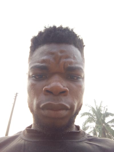 A close-up selfie shows a young Black man with a furrowed brow. He has short, dark, curly hair and a trimmed beard. His skin has a slightly oily sheen. He's wearing a dark, high-necked shirt. The background is an overcast sky. A utility pole is visible on the left, and a palm tree stands on the right, suggesting a tropical or subtropical environment. The lighting is bright and somewhat harsh.