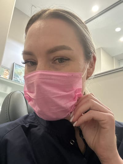 A person with light skin, wearing a pink surgical mask, is seen from a high angle. The person has blonde hair and is wearing a dark blue garment. They are adjusting the mask with their hand, which has long white fingernails. In the background, there are framed pictures on a wall and part of an office chair. The location is not identifiable as Kraskovo, Russia from the information given in the image.