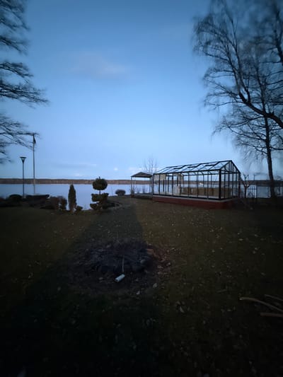The image depicts an outdoor scene in Baltezers, Latvia, at dusk. A glass greenhouse structure stands on a plot of land near a body of water, possibly a lake or a wide part of a river. The water is reflecting the sky. Bare trees are visible on either side of the frame. The ground is covered in grass and some dead leaves. To the right, near the edge of the frame, is some scattered debris, including what appears to be a white cup. A distant lamppost is seen across the water. The overall lighting is dim, suggesting the late hour.
