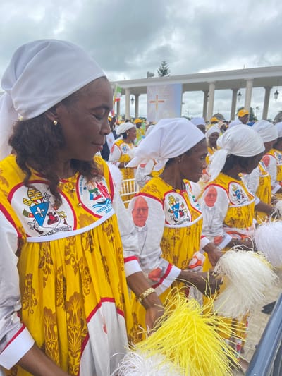 The image captures a group of people dressed in yellow and white garments, sitting outdoors with a building in the background. The individuals are wearing white head coverings and their attire features decorative emblems. Some are holding white or yellow objects resembling feather dusters. The setting appears to be an event or gathering taking place in an open area. It's not possible to determine the location or if it is in Mongomo, Equatorial Guinea, from the visual information alone.