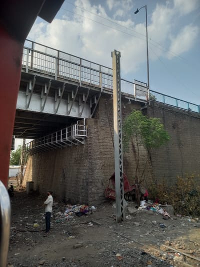 The image depicts a scene in Bhopal, India, dominated by an elevated structure, likely a railway overpass or flyover. The structure is constructed of stone and steel, with a stairway attached to its side. Debris and discarded items are scattered on the ground next to a person lying down. A streetlight and power lines are visible above. A tree grows out of the wall. A red vehicle is overturned.