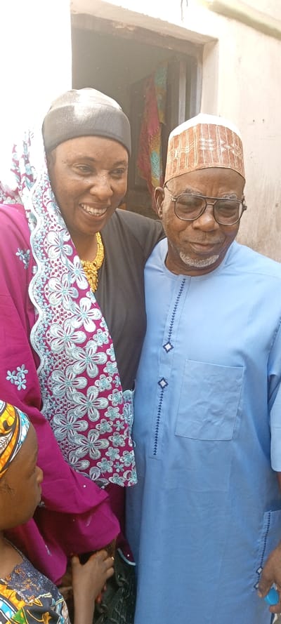 The image depicts three individuals in the city of Dan Maliki, Nigeria. An adult woman, positioned on the left, has dark skin and wears a black head covering and a black top. A magenta-colored garment with light blue floral embroidery is draped over her left shoulder and chest; she also wears a gold-colored necklace. She smiles broadly. Her left arm rests on the right shoulder of an older adult man beside her. The man, on the right, has dark skin, a white-grey beard, and wears dark-framed glasses. He is dressed in a light blue traditional tunic with dark blue geometric embroidery and a patterned light brown cap. His expression is a gentle smile, with eyes slightly downcast. Partially visible at the bottom left is a child, with dark skin, wearing a multi-colored patterned head wrap and top, facing left in profile. The man's right hand, visible at the bottom right, holds a small light blue cylindrical object. The background features a plain light-colored wall and a doorway with a dark interior where blurred, multi-colored fabrics are visible.