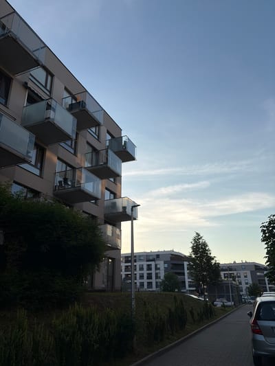 The image depicts an urban scene in Lublin, Poland. In the foreground, a modern apartment building with multiple balconies featuring glass railings is prominent on the left. The building's facade is a neutral beige color, and the balconies are cantilevered, creating a dynamic architectural profile. Lush green vegetation, including a dense hedge and tall grass, covers a sloping embankment in front of the building.

To the right of the apartment building, a paved walkway or sidewalk runs parallel to a road. Beyond the walkway, more apartment buildings are visible in the background. These structures have a contemporary design with white and grey facades and numerous windows. One of these buildings has a sign that reads "MEDICOM."

The sky above is a clear light blue with a few wispy clouds, suggesting it is either early morning or late afternoon. The lighting indicates a soft, natural illumination, with some golden hues from the setting or rising sun casting light on the building's upper floors and balconies.

A silver car is partially visible in the bottom right corner, suggesting it is parked on the street or the road. The overall atmosphere is one of a developed residential area within a city.
