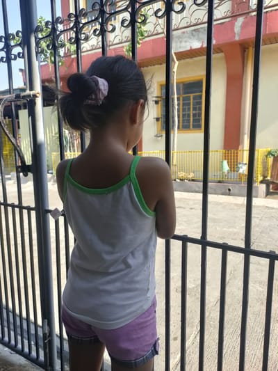 A young girl stands facing away from the camera, looking through a black metal gate. She has dark hair pulled back in a bun with a pink scrunchie. She is wearing a white tank top with green trim and purple shorts. The gate has ornate swirls at the top.
Behind the gate, a building with yellow and orange accents is visible. One window has a yellow frame. A yellow metal fence separates the yard from the street. The ground appears to be concrete. The sun is shining, suggesting it is daytime. The scene appears to be in a residential area. The girl seems to be observing something outside the gate, her posture suggesting a moment of quiet observation. There is no visible text in the image.