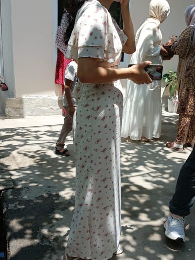 The image captures a group of people outdoors in what appears to be a city environment, possibly Osh, Kyrgyzstan, based on the hint. The setting is illuminated by sunlight, casting distinct shadows on the concrete ground, suggesting it is daytime.
In the foreground, a woman in a white floral print dress is prominently featured, her side facing the camera. She is holding a smartphone in her right hand, which is angled to display a picture on its screen. Her left hand is raised to her face, obscuring part of her features.
Behind her, another person, possibly a child, is visible wearing a white t-shirt and brown pants, with black sandals on their feet. Further back, a woman in a flowing white garment and a headscarf is seen holding a woven basket. To the right, another individual is wearing a patterned brown garment and sandals, and a third person is wearing dark pants and white athletic shoes.
The background shows a white building with some textured concrete elements and a doorway. A potted plant is visible to the right of the woman with the basket. The overall scene suggests a casual gathering or event taking place outdoors.