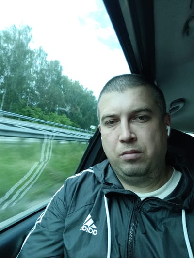 A close-up, slightly low-angle, selfie-style shot shows a man with short, dark hair and stubble on his face. He is wearing a dark grey hooded jacket with white stripes on the sleeves, and a white t-shirt underneath. A white earbud is visible in his right ear. The man is seated inside a vehicle, with the blurred background indicating motion. Through the car window, a highway can be seen with trees and greenery lining the side of the road. The sky appears overcast. The scene is consistent with a person traveling by car on a highway, possibly on the outskirts or approaching Třemošnice, Czechia, given the surrounding natural landscape.