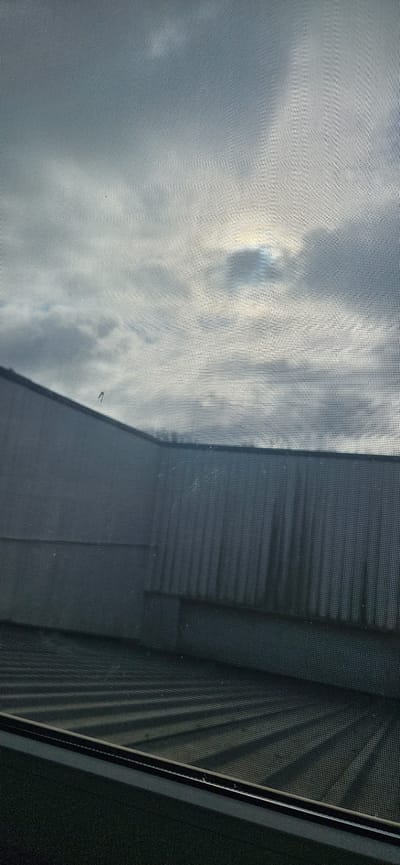The image shows a view through a window screen, with the sky and clouds visible on the left. The right side of the image shows a building exterior. The building appears to be made of corrugated metal or a similar material, with a repeating pattern of lines. The image is taken from inside a building, looking outwards. The location is in Delbrück, Germany, but there are no other people, interactions or activities captured in this image.