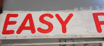This image shows a close-up of a sign painted with red letters on a white background. The letters spell out "EASY F". The sign appears to be weathered, with some of the white paint chipped and peeling, revealing glimpses of what looks like a grey surface underneath. There are faint horizontal lines drawn on the white background, possibly guidelines for the lettering. The overall impression is of an outdoor sign that has seen better days, possibly indicating a business or a service. The location context provided is Kipala Mpakani, Tanzania, which suggests this sign is situated in a community setting within that region.