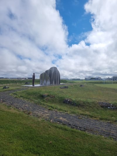 This is a vertically oriented, low-angle medium shot of a park-like area with a large, abstract metal sculpture and a lone, dark figure of a person standing to the left of the sculpture. The sculpture is comprised of several sleek, curved, metallic panels that rise from a paved surface. The figure, a dark silhouette, stands facing the sculpture.

The setting is an outdoor park with a grassy embankment on the right, leading up to a dark, brick-like pathway. A few dark rocks are scattered on the grass. In the background, a row of modern buildings is visible, suggesting a developed area. The sky is filled with dramatic white and grey clouds against a bright blue sky, indicating a day with variable weather. The lighting suggests it is daytime.

The scene conveys a sense of quiet contemplation, with the solitary figure and the large, imposing sculpture. The overall mood is somewhat serene and perhaps a bit melancholic due to the lone figure. There is no visible text in the image. The location is Sandgerði, Iceland.