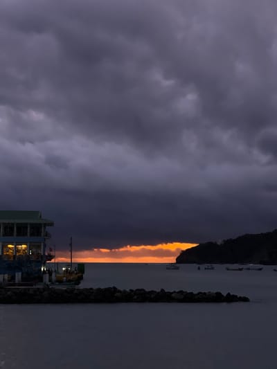 This is a photograph of the bay of San Juan del Sur, Nicaragua at dusk. The sky is filled with dark, dramatic storm clouds, with a sliver of orange and yellow light visible near the horizon, indicating the setting sun. In the foreground, a breakwater made of large, dark rocks juts out into the water. To the left, a multi-story building with a green roof and bright interior lights stands on a dock. Several small boats are anchored in the bay. The water is calm and dark.