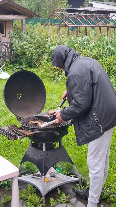 A person wearing a black hooded jacket and gray sweatpants is grilling food on a black charcoal grill in a grassy yard. The grill is open, revealing meat on the grates. The person is holding tongs and a black grilling tool, appearing to turn or move the food. In the background, there is a wooden structure resembling a small shed or outbuilding, a greenhouse with a metal frame, and lush green vegetation. The ground appears wet, suggesting recent rain. The scene is set in a rural or semi-rural outdoor environment.