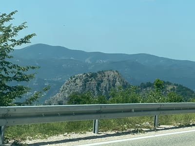 The image captures a mountainous landscape in Kućišta, Montenegro, viewed from a roadside. In the foreground, a metal guardrail runs horizontally across the frame, separating the road from the embankment. The embankment is covered with green grass, gravel, and some scattered white stones. Behind the guardrail, lush green trees and bushes are visible, with a prominent rocky outcrop in the middle ground. Further in the background, layered mountain ranges fade into a hazy blue sky. A small black speck, possibly a bird or insect, is visible in the upper left portion of the sky. The lighting suggests it is daytime, likely a clear or partly cloudy day.