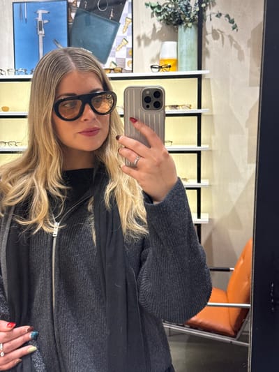 A woman with blonde hair and black glasses poses, holding a silver phone. She wears a gray sweater and a black scarf or jacket. The background features a mirror reflecting a room with shelves displaying eyeglasses, decorative plants, and an orange chair. The phone's case is metallic, and the woman wears a ring. Her nails are painted in different colors on each hand.